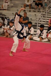 Young girl in a white dobok with a blue belt performs a martial arts pose on a pink mat, raising her right arm as spectators watch from the bleachers in the background