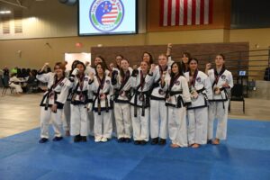 Group of smiling martial artists in white uniforms with medals posing on blue mats indoors, raising fists in celebration.