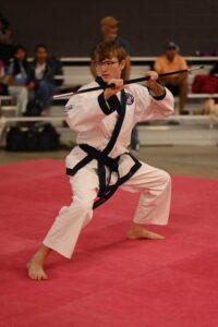 Young martial artist in a white dobok with a black belt practicing a staff form on a red mat, audience in the background.