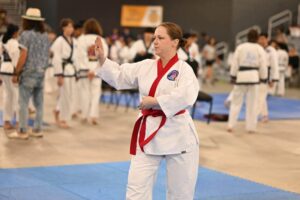 Female martial artist in a white karate gi with a red belt raises a blocking hand during a tournament with other competitors in the background.