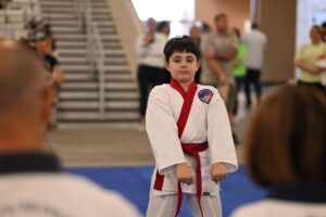 Young boy in a white martial arts gi with a red belt performing a stance in a gym, badge with the American flag on his chest, blurred spectators in the background.
