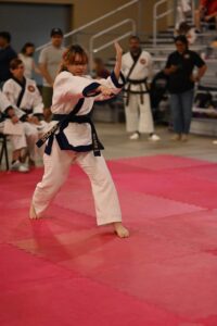 Young girl in a white karate gi with a navy belt performs a punch on a pink mat, with spectators in the background.