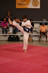 Young martial artist in a white gi performing a high kick on a red competition mat, with spectators in the background.