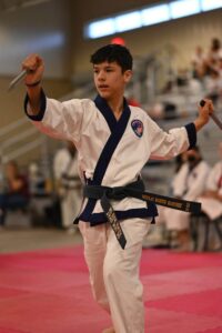 Young martial artist in a white gi with blue trim performing a weapon demonstration in a gym, holding a blade in each hand with arms extended.