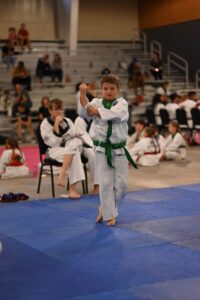 Boy in a white gi with a green belt performs a punch on blue mats at a martial arts event, spectators in the background.