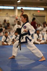 Female taekwondo practitioner in a white gi with a blue belt performs a high kick on blue mats, spectators in the background.