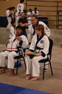 Young taekwondo student and female teammate seated in chairs wearing white uniforms; coach leans in behind them during practice in a gym, with other students in the background.