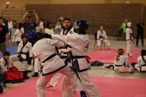 Two martial artists sparring in white uniforms on pink mats, wearing headgear and gloves, indoors with spectators in the background.