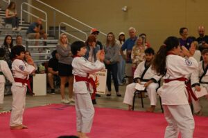 Young martial artists in white gi and red belts practicing on a pink mat as spectators watch from the bleachers behind them on a gymnasium floor.